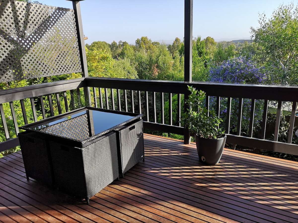 An outdoor balcony space is presented with wooden decking and a stylish black rattan coffee table. Lush greenery is visible in the background, enhancing the serene atmosphere. A potted plant adds a touch of nature, making the space inviting for relaxation.