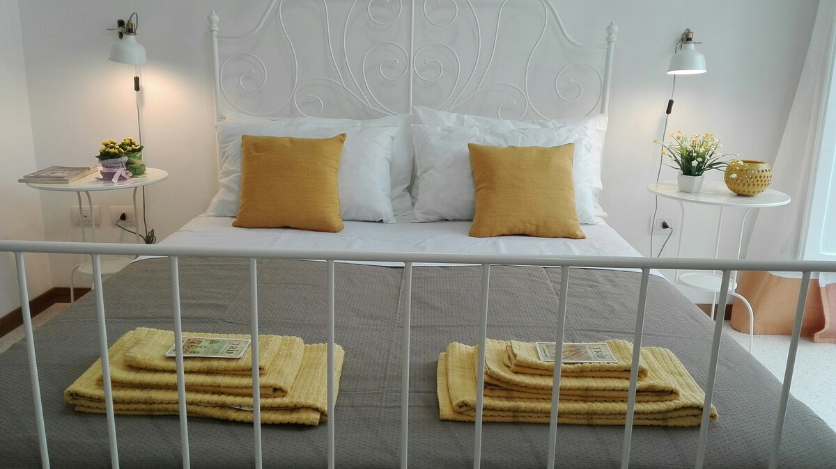 A spacious bed with a white iron frame is complemented by soft, white linens and two yellow decorative pillows. Neatly folded yellow towels are arranged on the bed, while bedside tables with lamps and small potted plants add a touch of tranquility to the setting.