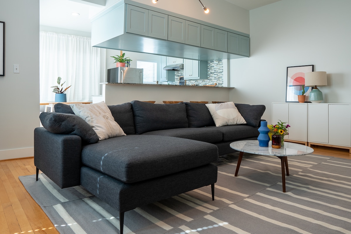 A modern living area features a dark sectional sofa with plush cushions and a light-colored area rug with geometric patterns. A round coffee table sits in front, adorned with a small vase of flowers. A kitchen area is visible in the background with sleek cabinetry and stone accents.