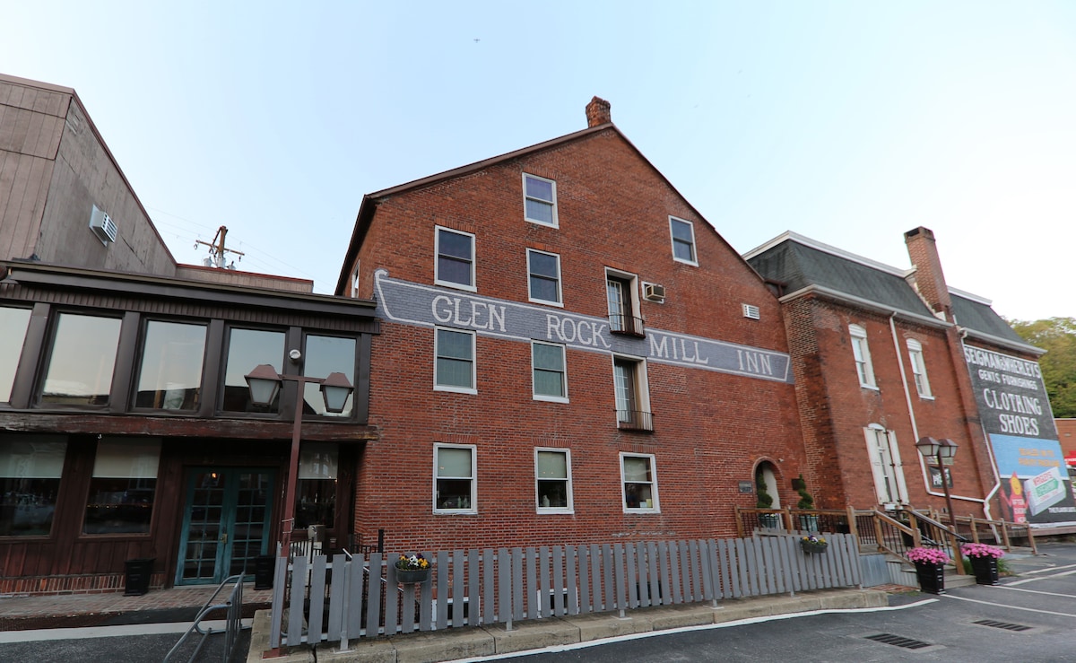 The exterior of the Glen Rock Mill Inn is showcased, featuring a historic red brick facade with large windows. A sign indicating the inn's name is prominently displayed. The building is flanked by additional structures, and flower planters are placed along the front.