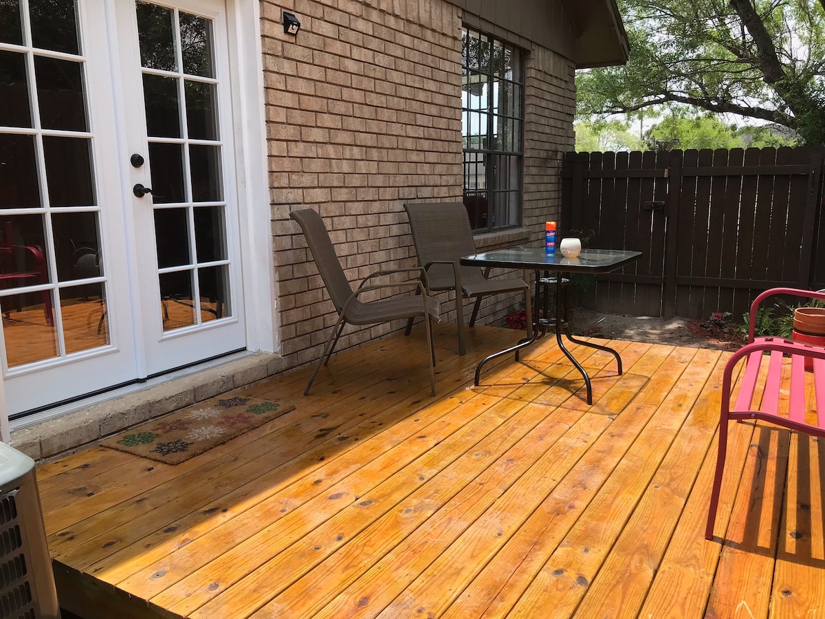 A wooden deck area is shown with two metal chairs and a glass table at the center. French doors lead into the house, illuminating the space. A pop of color is provided by a pink chair nearby, while greenery is visible in the background.