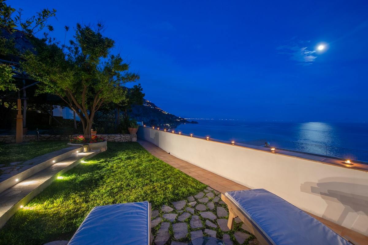 The garden area is illuminated softly at night, with a moonlit view of the sea. Two sun loungers are positioned on the grassy lawn, surrounded by stone pathways and potted plants, creating a tranquil outdoor space for relaxation.