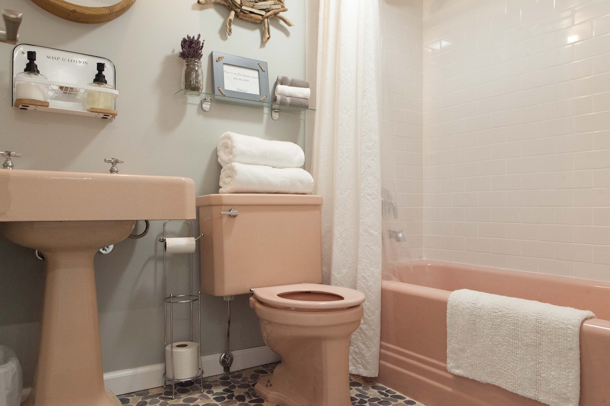 The bathroom features a pastel pink color scheme with a ceramic sink and toilet, complemented by soft, plush towels stacked neatly on a shelf. A tub with a shower curtain stands next to the tub, and a pebble tile floor adds texture to the space.