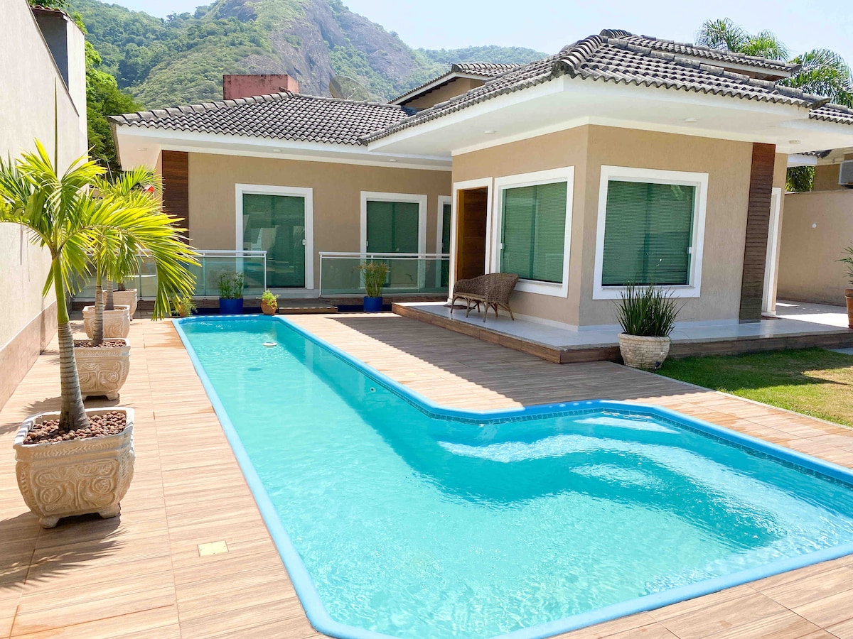 An outdoor area features a clear blue swimming pool surrounded by sunlit tiles and potted plants. The modern house is positioned to showcase large windows that allow natural light to fill the interior. Lush green hills are visible in the background.