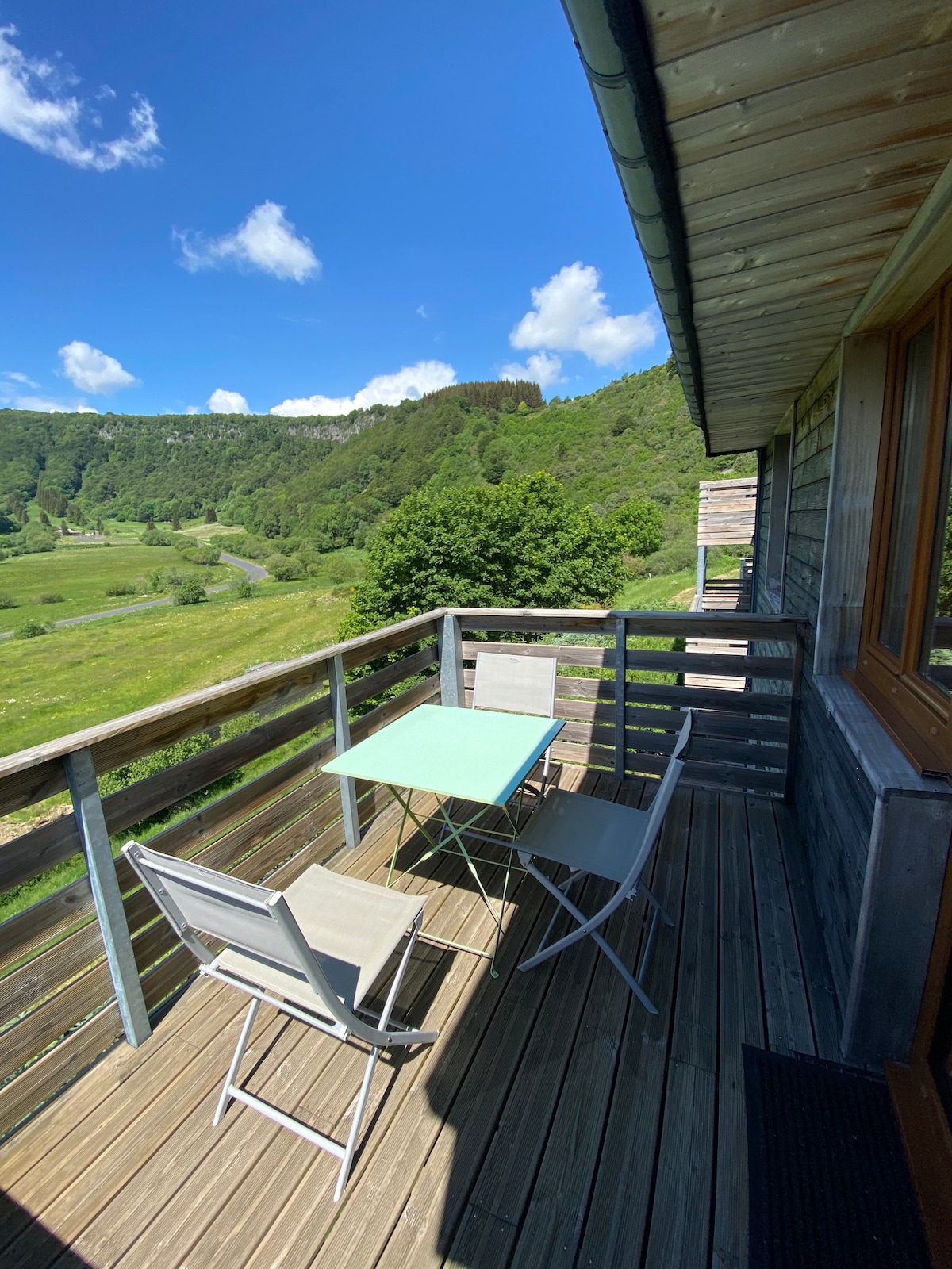 A small balcony is shown with a light blue table and two chairs, surrounded by wooden railings. Lush green hills and a clear blue sky create a serene backdrop, while sunlight casts gentle shadows on the deck.
