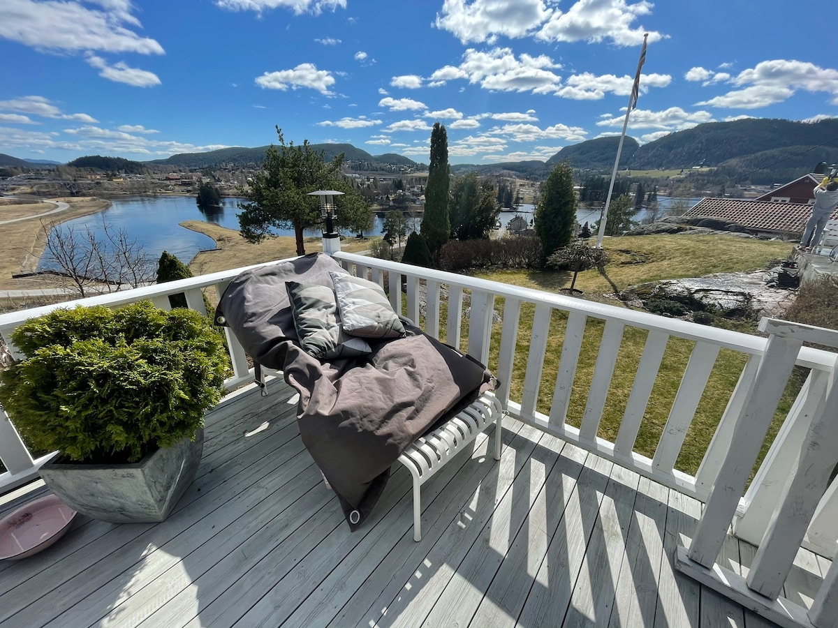 A patio area is shown with a comfortable lounge chair adorned with soft pillows. Lush greenery is situated in a nearby planter. The scene reveals expansive views of the surrounding landscape and water, under a clear blue sky with scattered clouds.