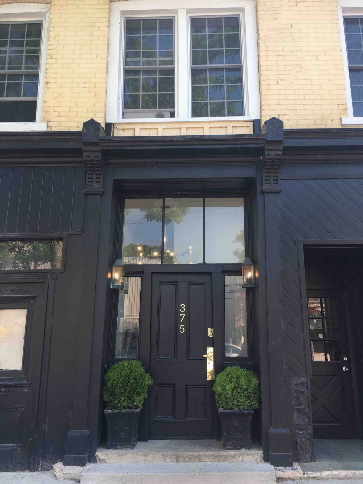 The exterior features a striking black facade accentuated by tall windows and ornate detailing. The entrance is framed by symmetrical potted plants, while the door is marked with the number 375. A warm glow emanates from the wall sconces on either side.