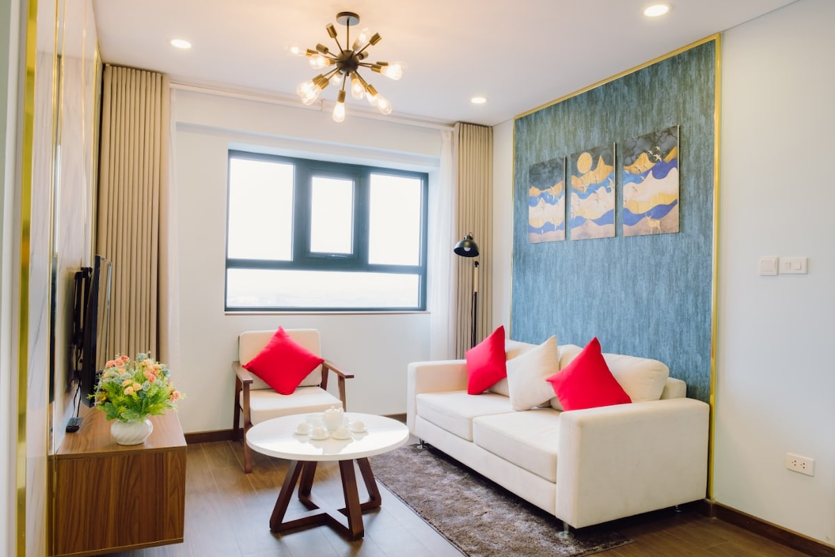 A cozy living area is furnished with a light-colored sofa adorned with red cushions. A round coffee table is positioned in front, with a floral arrangement nearby. Natural light enters through a large window, and a modern light fixture hangs from the ceiling.