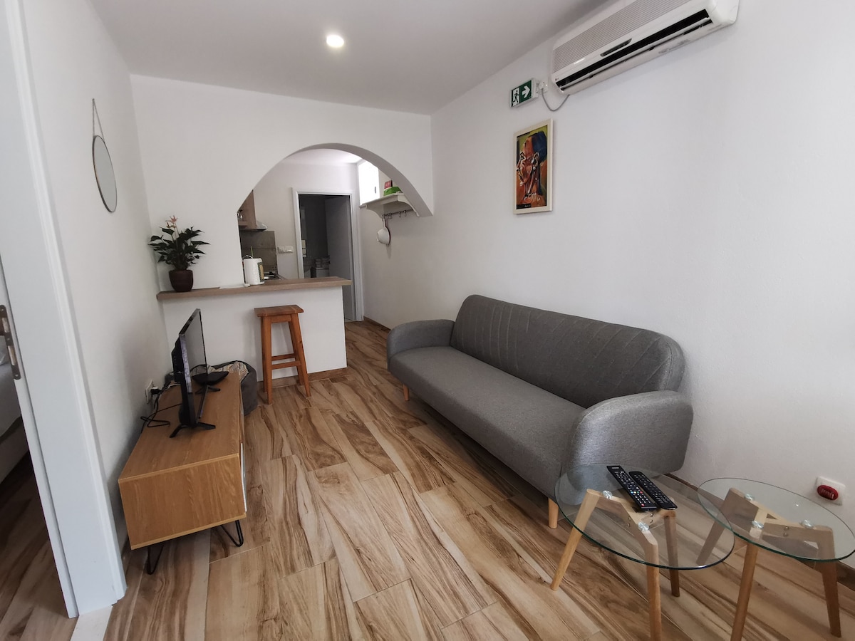 A cozy living area is presented with a light gray sofa and a small glass coffee table with wooden legs. A television sits on a wooden stand, and a small wooden stool is positioned nearby. An archway leads to a well-lit kitchen area in the background.
