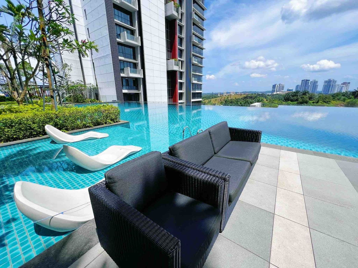 A modern outdoor lounging area features dark wicker seating facing an infinity pool. The pool's clear water reflects the sky, and lounge chairs are positioned nearby. Lush greenery surrounds the space, with a city skyline visible in the distance.