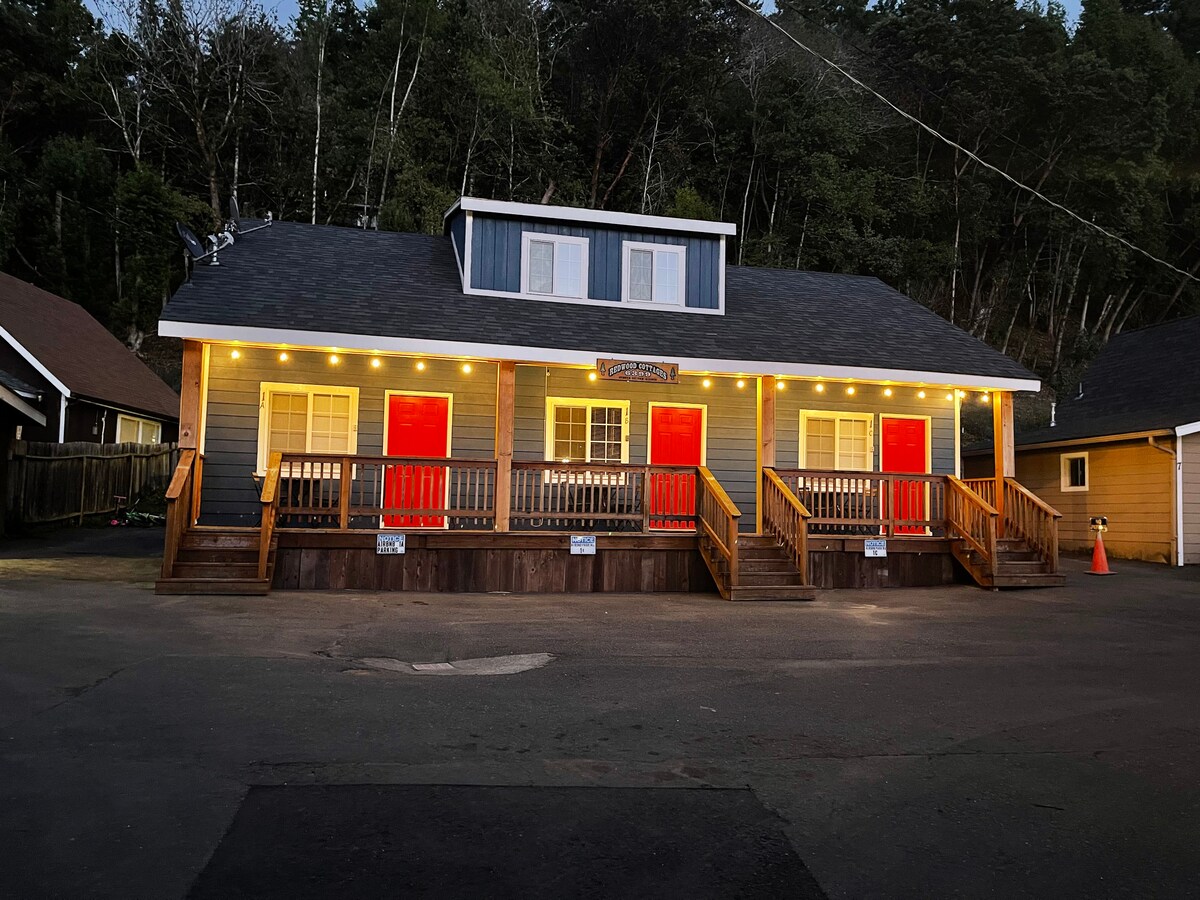The exterior of the building is shown, featuring a two-story structure with a welcoming design. Bright red doors are framed by warm lighting along the facade. A wooden deck with steps leads to the entrance, surrounded by trees, creating a serene atmosphere.
