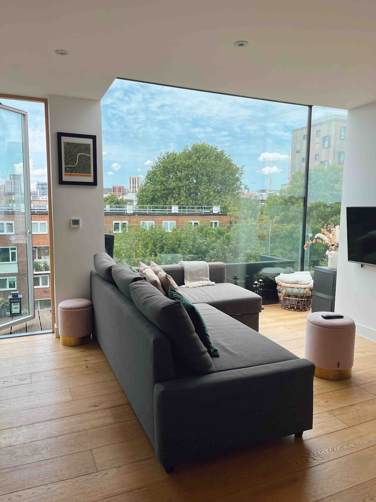 A bright living area is framed by large glass windows that provide a view of the surrounding greenery and cityscape. A comfortable gray sofa is positioned next to a light-colored ottoman. Soft natural light enhances the warm wooden flooring throughout the space.