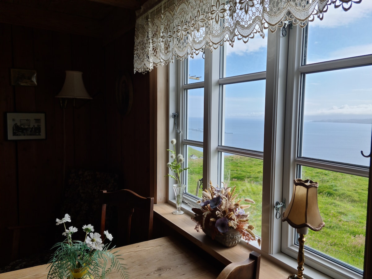 A dining area features large windows with lace curtains, providing a view of the peaceful surroundings and sea. A wooden table is set with floral decorations, while a small lamp sits beside a vase filled with shells, adding a touch of charm to the space.