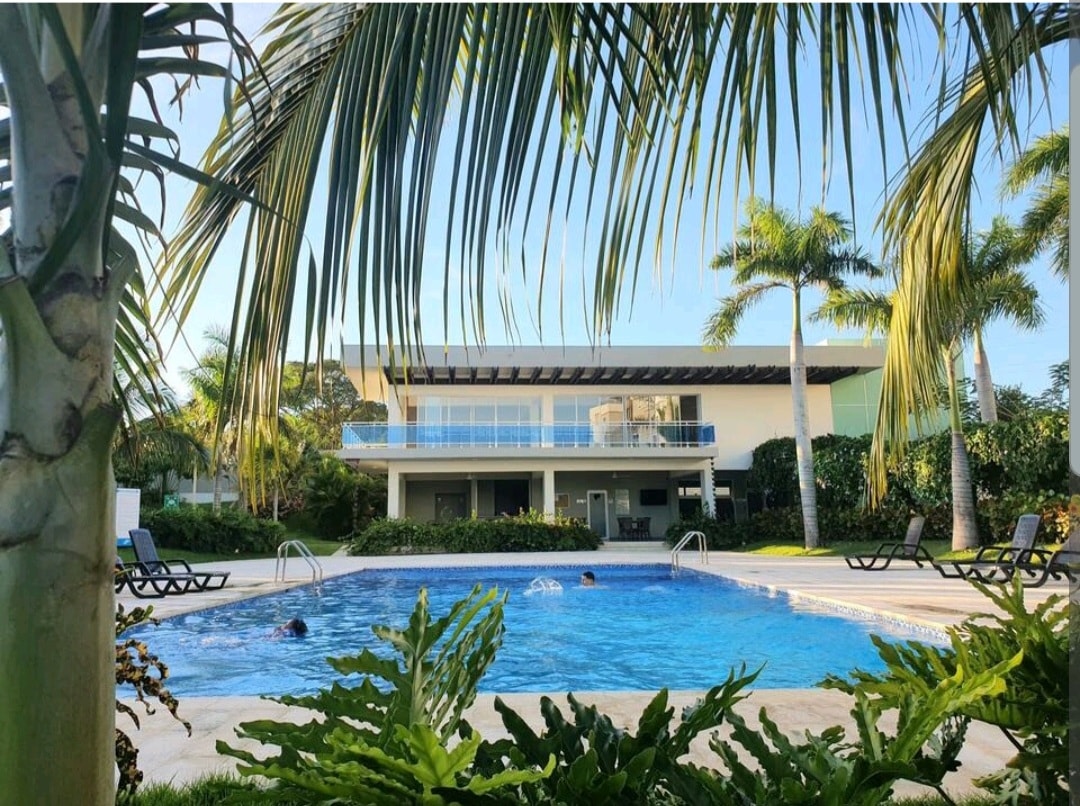 A large pool is surrounded by tropical greenery and lounge chairs. The spacious building is visible in the background, featuring a covered balcony and large glass windows that enhance the outdoor experience.