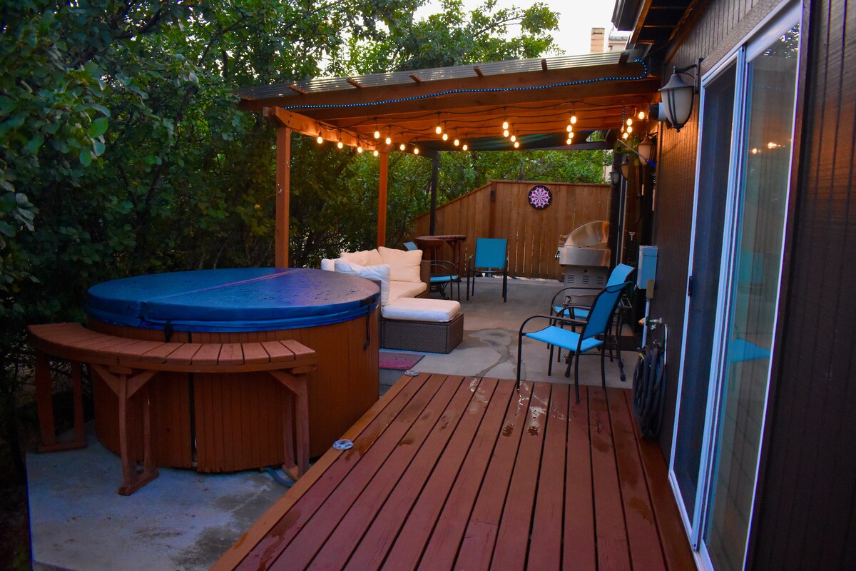 A private outdoor space features a hot tub surrounded by greenery and soft lighting. Comfortable seating, including a sofa and chairs, provides options for relaxation, while a wooden deck offers a welcoming atmosphere for gatherings.