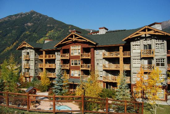 The building, featuring a blend of wood and stone materials, rises three stories amidst landscaped greenery. Balconies with wooden railings provide views of the surrounding mountains. A pool area is visible in the foreground, alongside tall trees adorned with golden autumn leaves.