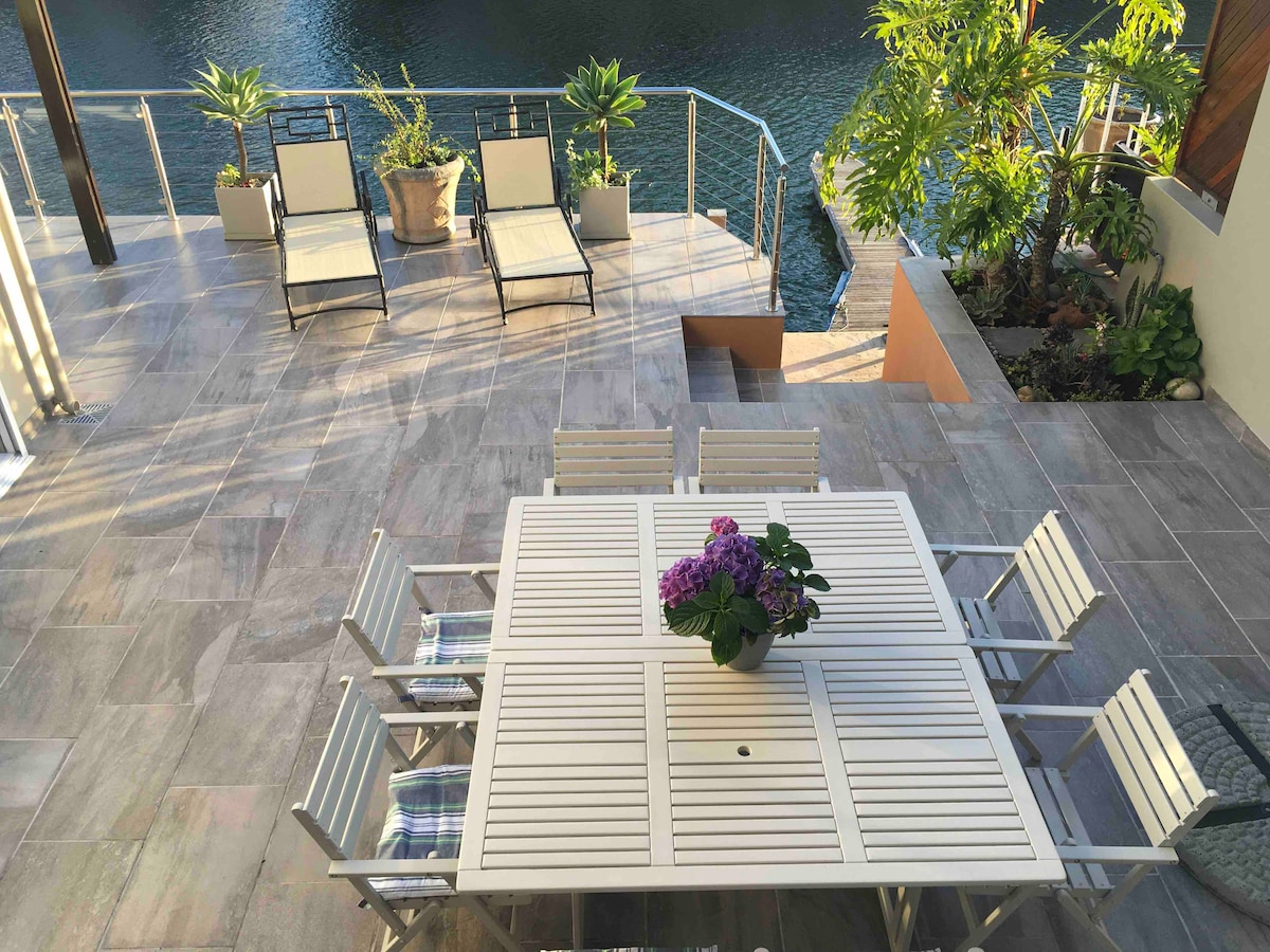 An outdoor patio area features a large white dining table surrounded by eight matching chairs, complemented by a potted plant with colorful blooms. Sun loungers are positioned nearby, offering views of the water, while greenery enhances the peaceful environment.