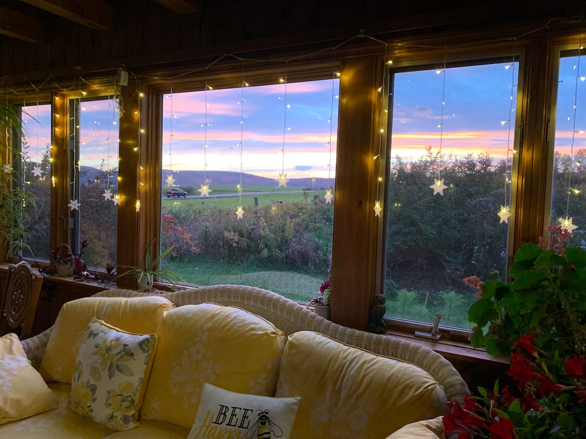 A sunroom with large windows showcases a vibrant sunset, framed by twinkling fairy lights. A comfortable yellow sofa with floral accents invites relaxation while potted plants add a touch of nature to the serene countryside view outside.