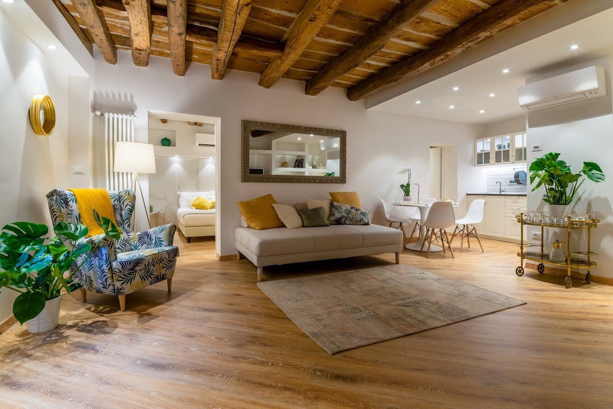 The spacious living area features wooden beams on the ceiling, enhancing the historic charm. A patterned armchair and a neutral sofa are complemented by a coffee table and indoor plants. Dining chairs surround a white table, while the well-equipped kitchen area is visible in the background.
