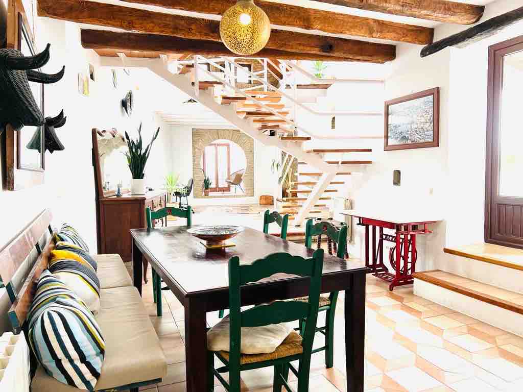 An open dining area displays a large wooden table surrounded by colorful, green chairs. Natural light filters through a nearby window, illuminating the warm tiles and wooden beams overhead. A modern staircase leads to the upper levels, adding depth to the inviting communal space.