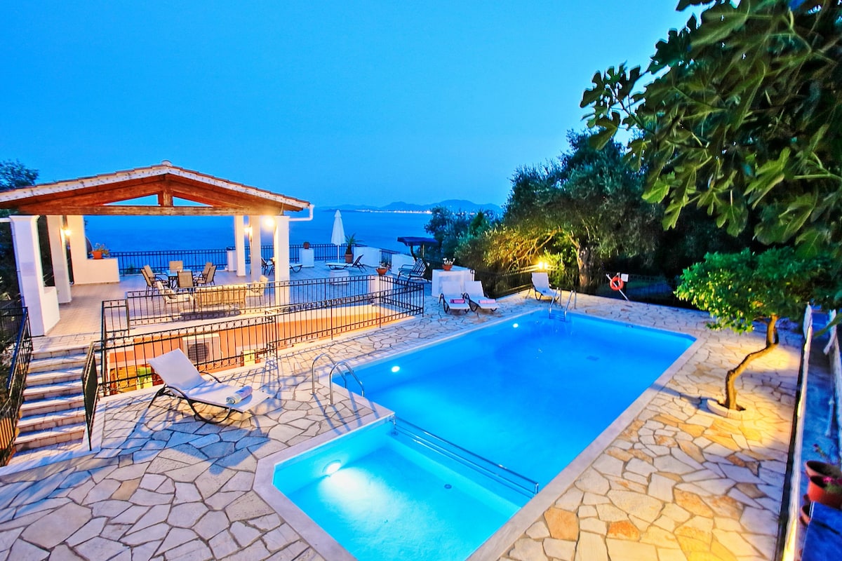 The outdoor terrace is complemented by a private swimming pool, illuminated in soft blue lights. Comfortable loungers surround the pool area, and a shaded seating area provides a relaxing space with panoramic views of the Ionian Sea.