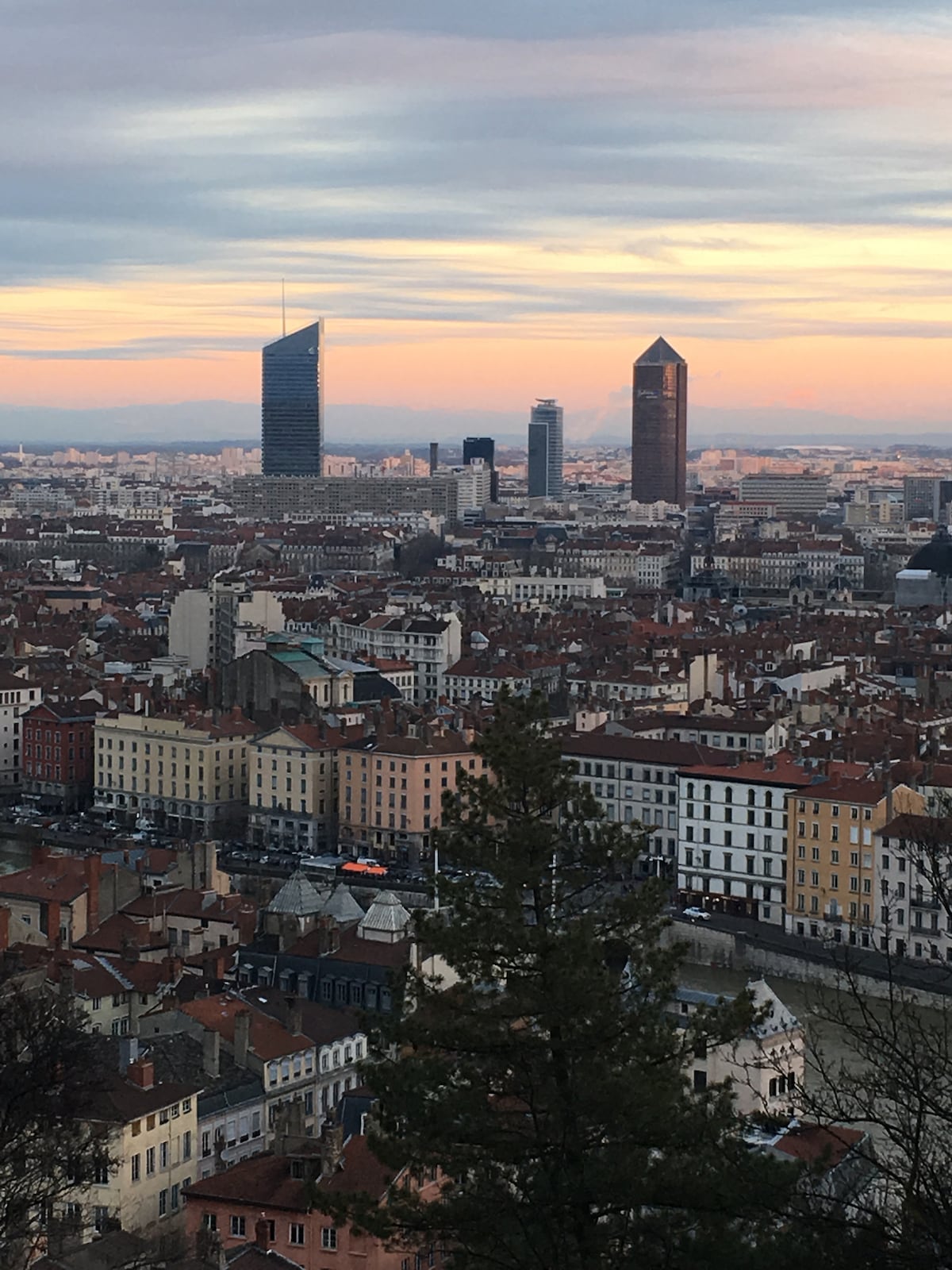 A panoramic view of Lyon showcases a mix of historic and modern architecture. The skyline is dominated by contemporary towers rising above traditional rooftops, with the Saône river flowing in the foreground, reflecting the soft hues of the sunset in the sky.