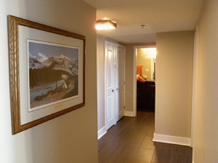 A well-lit hallway features modern flooring and neutral walls. A framed landscape print hangs to the left, while doors lead to rooms at the end. Recessed lighting above provides illumination, enhancing the sense of space as the entrance to the suite is approached.