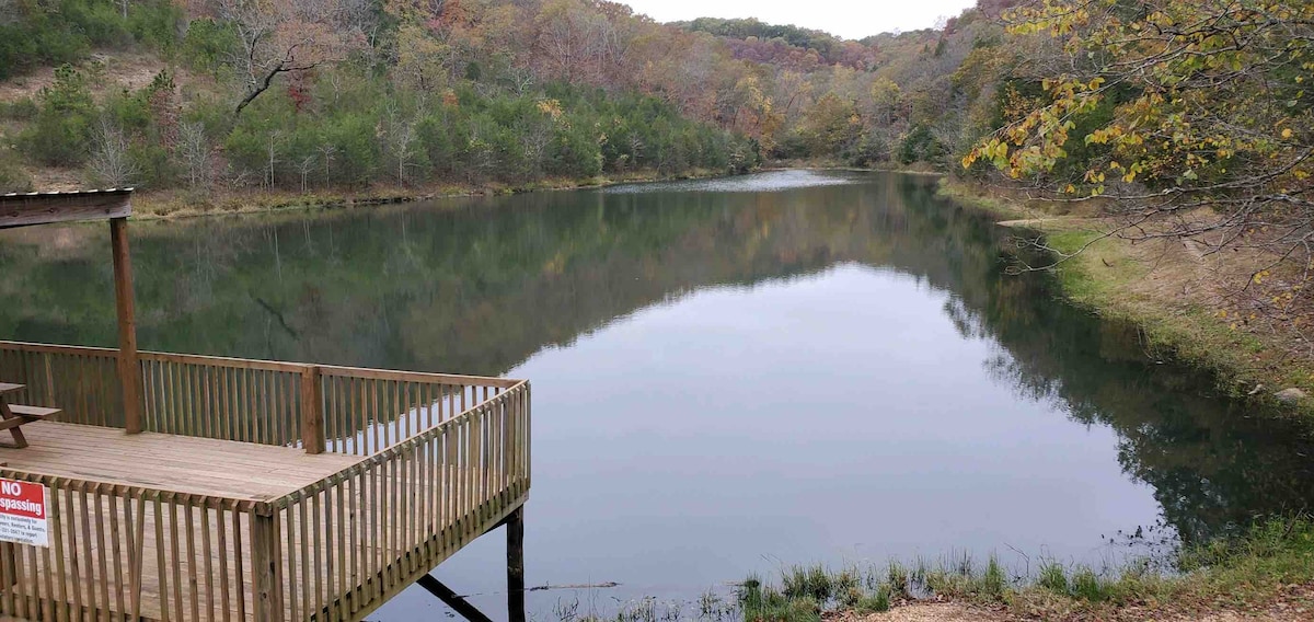 A serene fishing pond is framed by lush greenery and autumn-colored trees. A wooden deck extends over the water, providing a peaceful spot to relax and enjoy the natural surroundings, with calm reflections on the pond's surface beneath a partially cloudy sky.