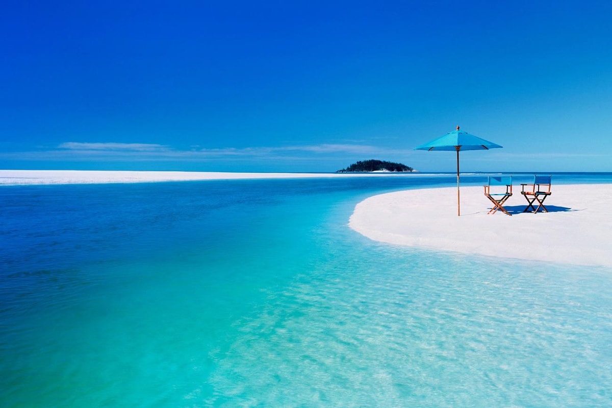 A serene beach scene features soft white sand meeting clear turquoise waters. An umbrella provides shade for two beach chairs positioned on the shore. The horizon showcases the calm sea, blending into a bright blue sky, creating a peaceful coastal environment.
