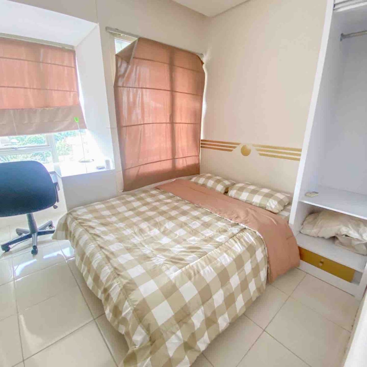 A cozy sleeping area with a queen bed is dressed in a checked beige and white duvet. Natural light fills the space through partially open brown curtains. A simple chair and a wardrobe can be seen, offering essentials for a comfortable stay.