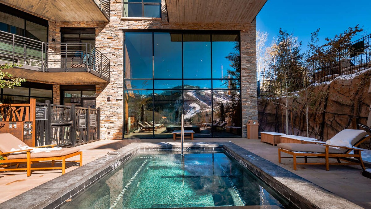 An outdoor spa pool is surrounded by stone walls and features wooden lounge chairs. Floor-to-ceiling glass windows provide views of the mountain landscape. The area is well-lit, highlighting the inviting water and tranquil ambiance beneath the clear sky.