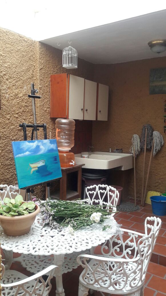 A charming outdoor space features a white wrought-iron table surrounded by matching chairs. A potted plant sits atop the table, while an easel holds a colorful painting. Cabinets and a water dispenser are visible, alongside a sink and cleaning tools, all set against a textured earth-toned wall.