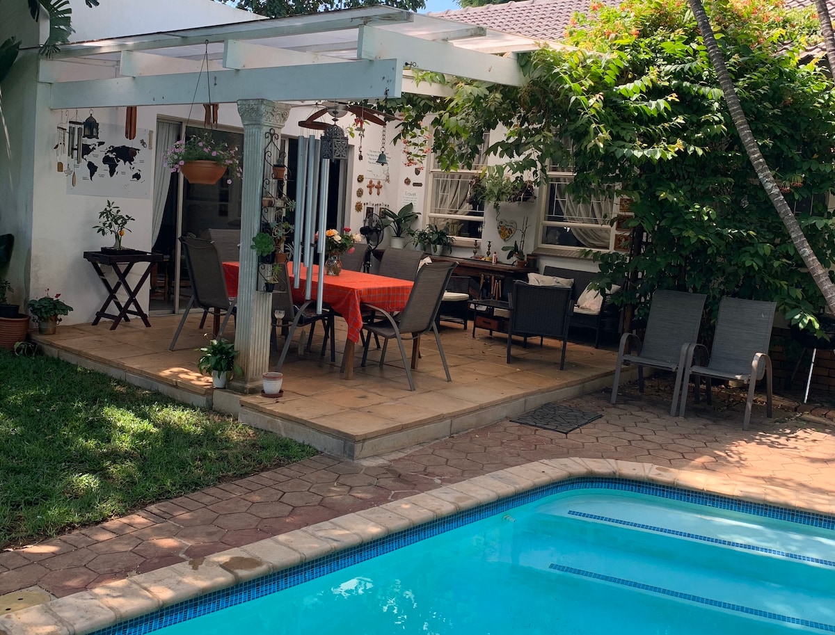 An outdoor patio is featured, showcasing a dining table with an orange tablecloth and several chairs. Surrounding greenery provides a natural ambiance, while a swimming pool glimmers nearby. Relaxation spots include sun loungers positioned on the stone-paved area next to the pool.