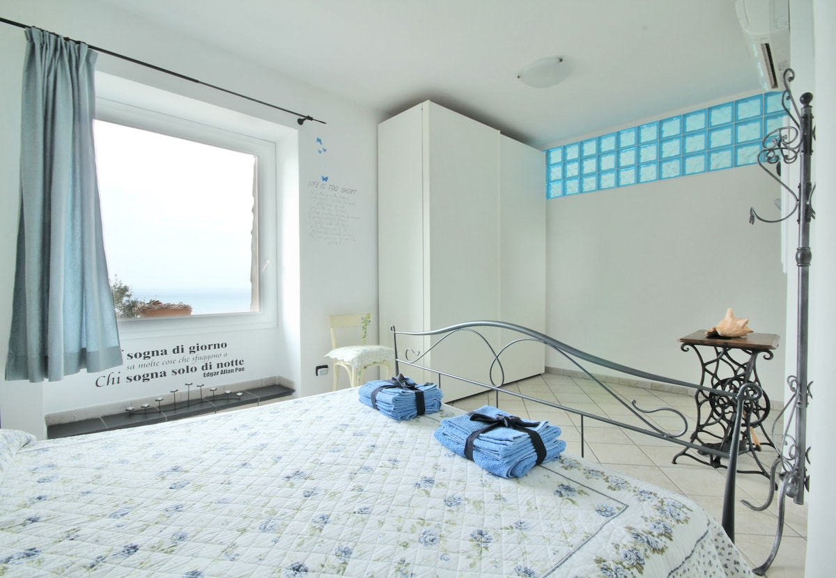 A spacious sleeping area features a metal bed frame with a light floral-patterned bedspread. Two neatly rolled towels are placed on the bed. A large window offers a view of the sea, complemented by light blue accents on the walls and curtains.