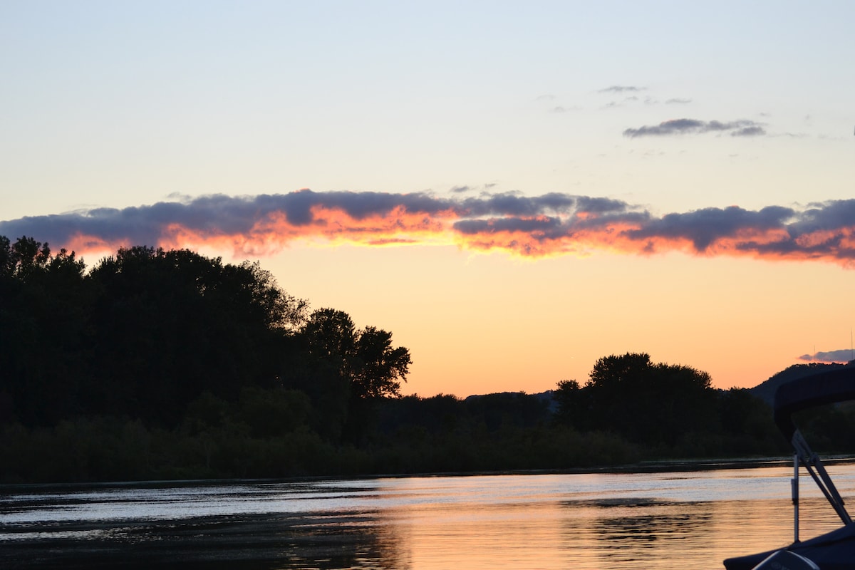 A sunset casts vibrant colors across the sky, with clouds illuminated in shades of orange and pink. The stillness of the water reflects the hues, while silhouettes of trees line the riverbank, creating a serene evening scene.