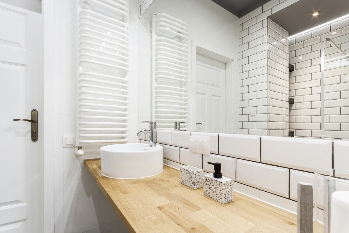 A modern bathroom features a wooden countertop supporting a round washbasin. White tiles cover the walls, complemented by a heated towel rail. A large mirror extends across the sink area, enhancing the bright and clean aesthetic of the space.