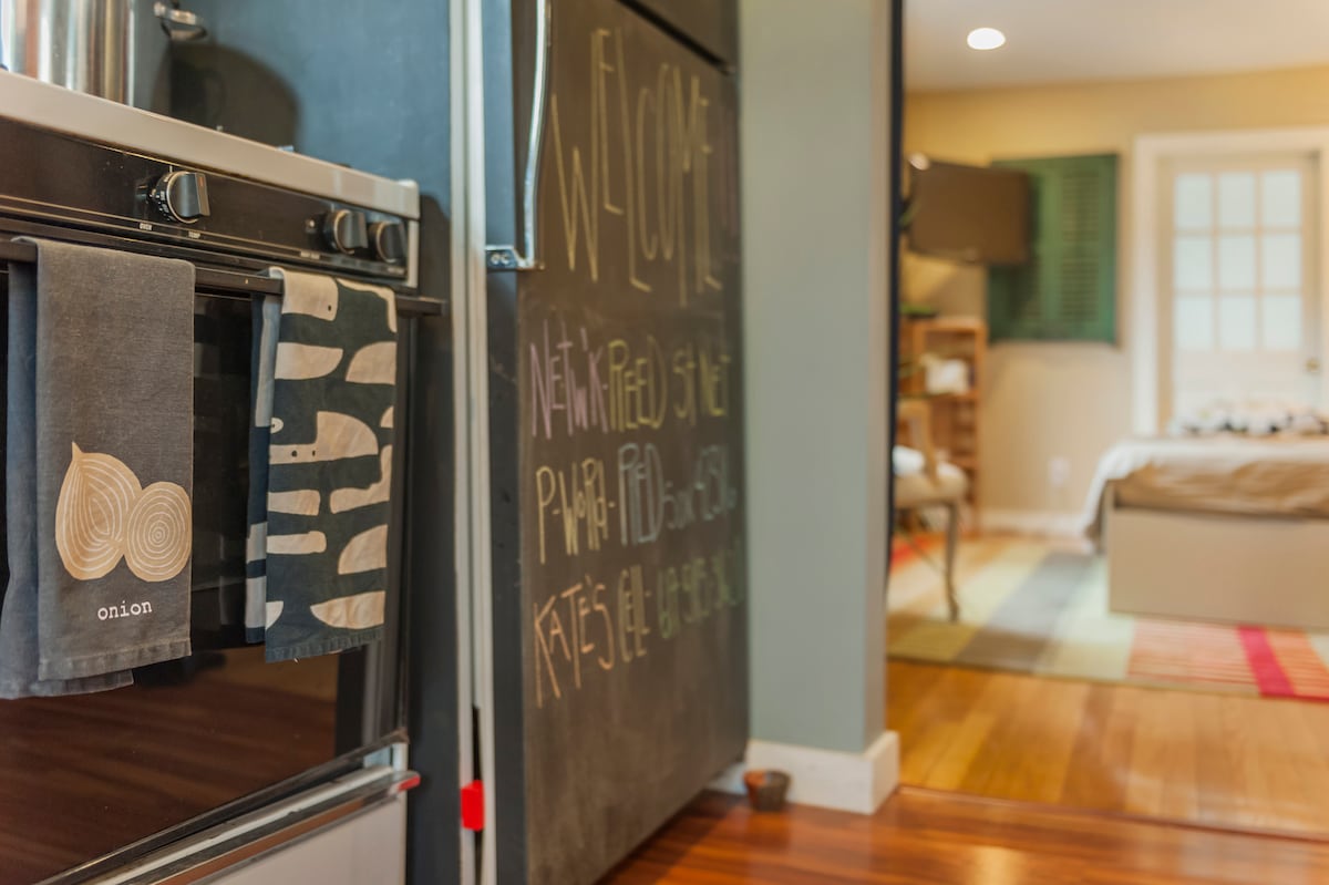 A welcoming kitchen area is captured, showcasing a modern gas stove with neatly arranged towels. A chalkboard displays a warm greeting, while the doorway leads to a well-lit living space with wooden flooring and a cozy atmosphere.
