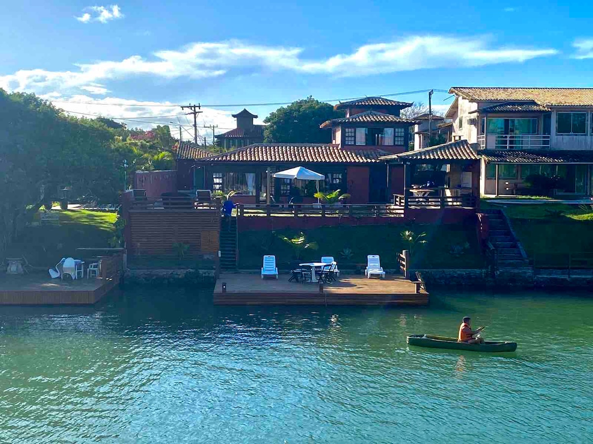 A view of the property from the water, showcasing the two-story house with a tiled roof and wooden deck. Lounge chairs are arranged on the deck overlooking the canal. A person is seen kayaking in the calm waters, with lush greenery framing the scene.