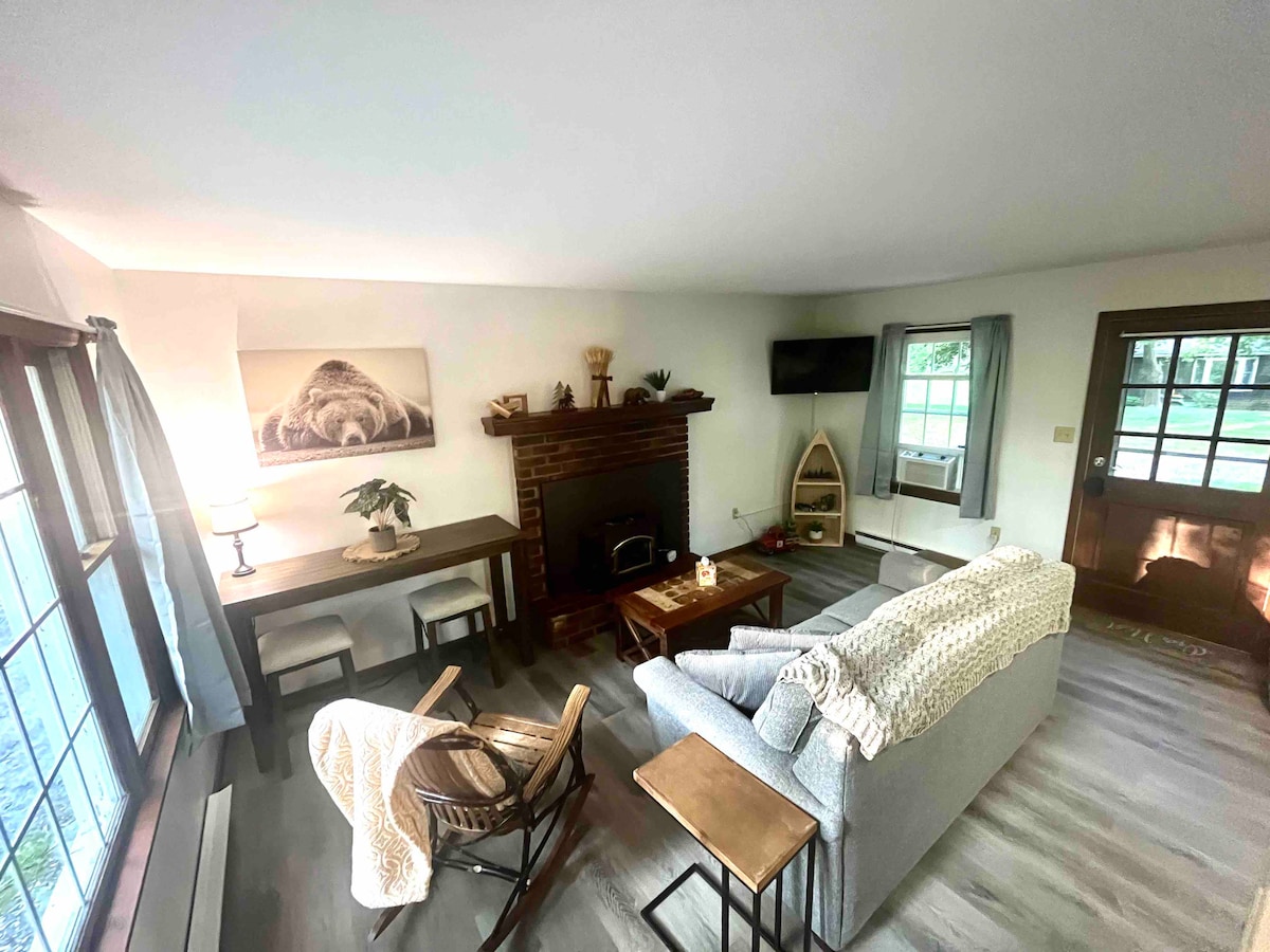 A well-lit living area is presented, featuring a cozy grey sofa and wooden coffee table. A small wooden desk sits adjacent to a decorative plant. A stone fireplace is visible, complemented by a wall-mounted television and natural light streaming in through the large windows.