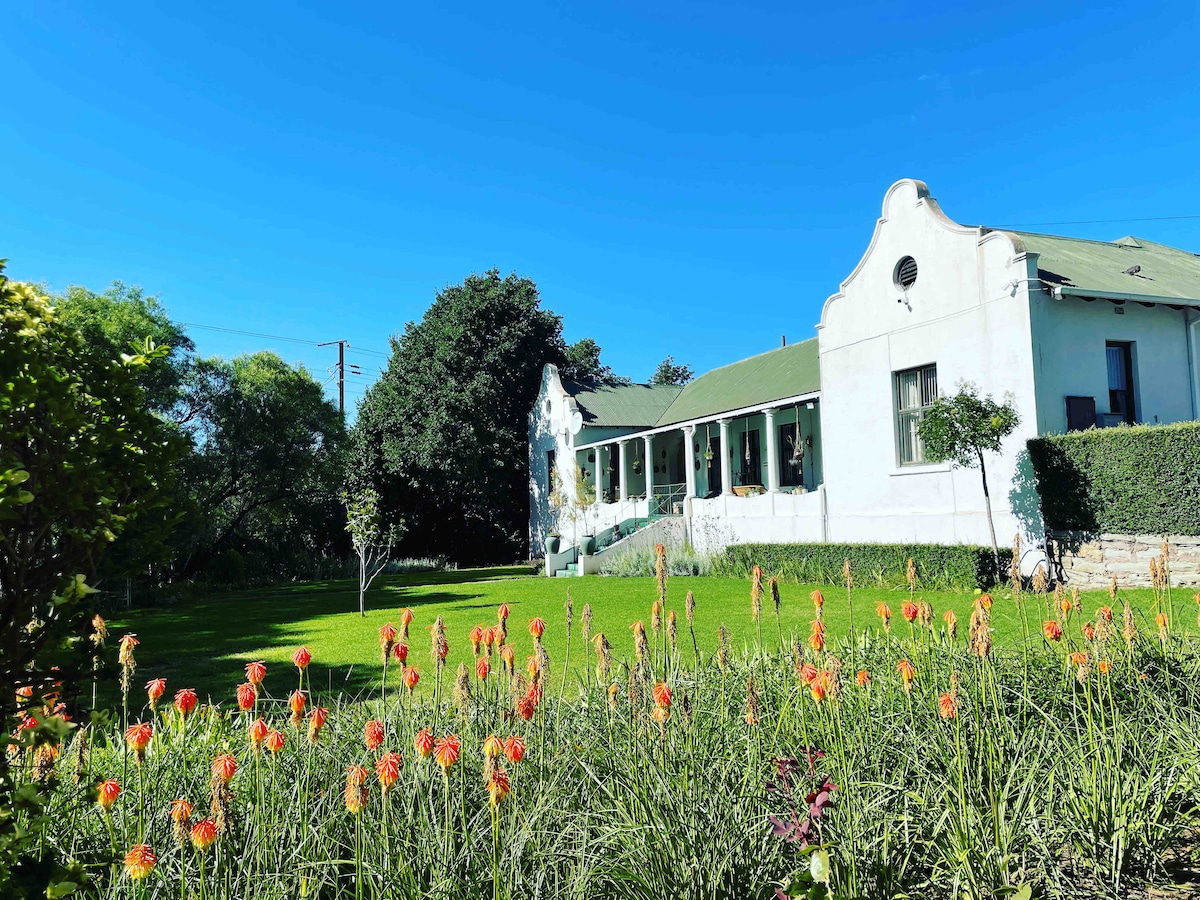 The historic house is showcased against a clear blue sky, featuring a green roof and a long row of large windows. Lush greenery surrounds the property, with vibrant orange flowers blooming in the foreground, creating a welcoming entrance to the garden area.