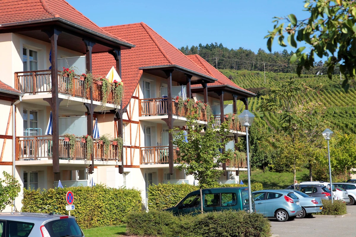 A charming residence is depicted with multiple levels, featuring balconies adorned with flowerboxes. Lush greenery surrounds the building, and rows of vineyards are visible in the background, providing a scenic view. Parking is available at the forefront, with several cars positioned nearby.