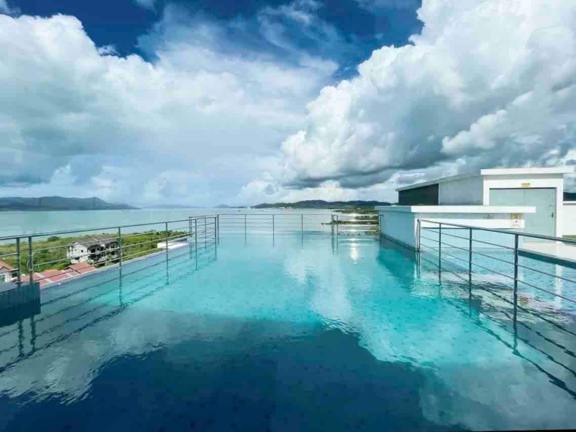 An elevated sky pool reflects the surrounding clouds and sky, offering a view of the distant mountains and coastline. The pool's water appears still and inviting, framed by sleek railings, while the landscape extends beyond, enhancing the sense of tranquility.