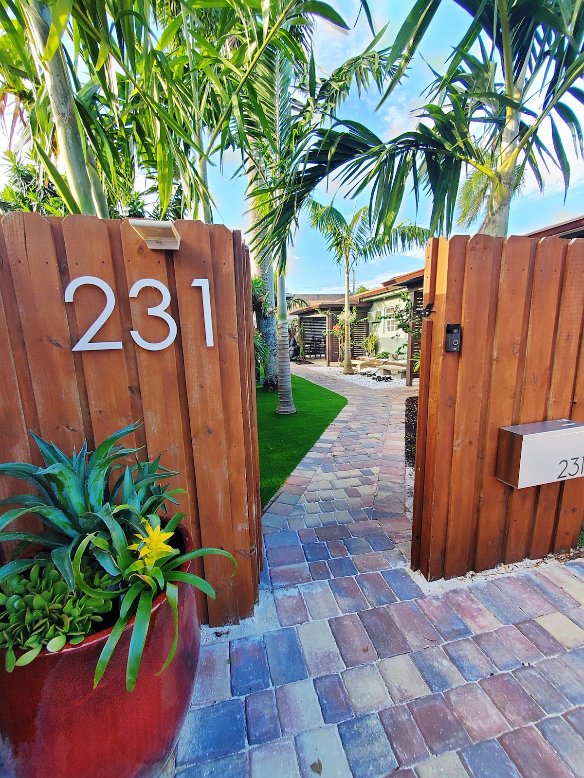 An arched wooden gate displays the number '231' and opens to a stone pathway lined with palm trees. Lush greenery and a vibrant flowerpot add a welcoming touch, leading to a courtyard area in the distance.