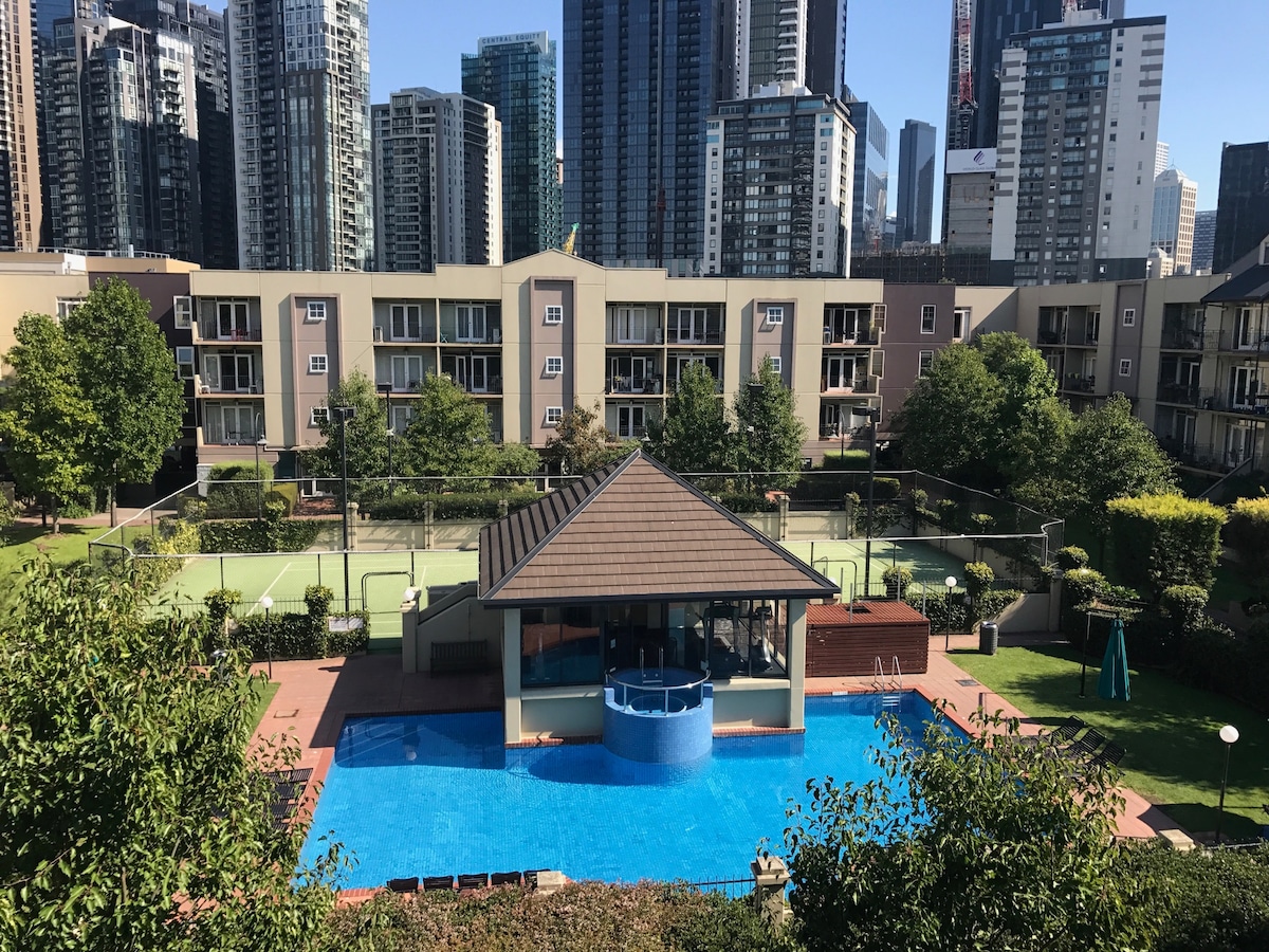 An outdoor area is depicted, featuring a bright blue solar-heated pool surrounded by landscaped greenery and buildings. A grass tennis court is visible nearby, with a sheltered seating area adjacent to the pool, creating a relaxing environment within the complex.