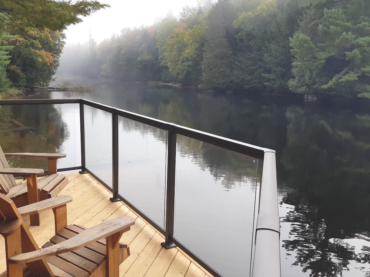 A wooden deck features two comfortable chairs overlooking a serene river. Mist covers the water's surface, while trees with varying shades of green and hints of autumn colors frame the scene, creating a peaceful and private atmosphere.