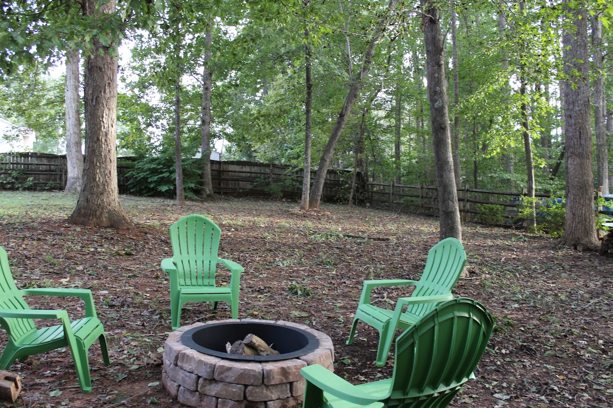 A cozy backyard setting features a stone fire pit surrounded by four green Adirondack chairs. Tall trees provide shade, while a wooden fence offers privacy in the background. The ground is covered with fallen leaves, creating a natural, inviting atmosphere.