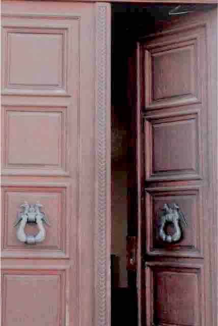 The entrance features large wooden doors with intricate paneling, partially opened to reveal a glimpse of the interior. Brass door knockers are mounted on each door, adding a touch of elegance to the inviting entryway.