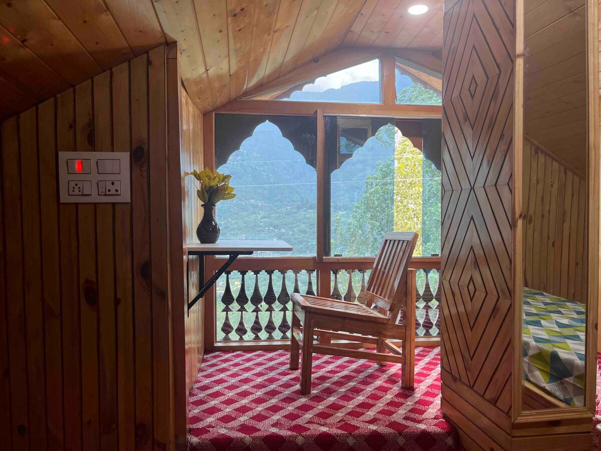 A cozy nook features a wooden chair and a small table, inviting relaxation while surrounded by large windows showcasing stunning mountain views. The red and white patterned carpet adds warmth to the wooden interior, enhancing the peaceful ambiance of the space.