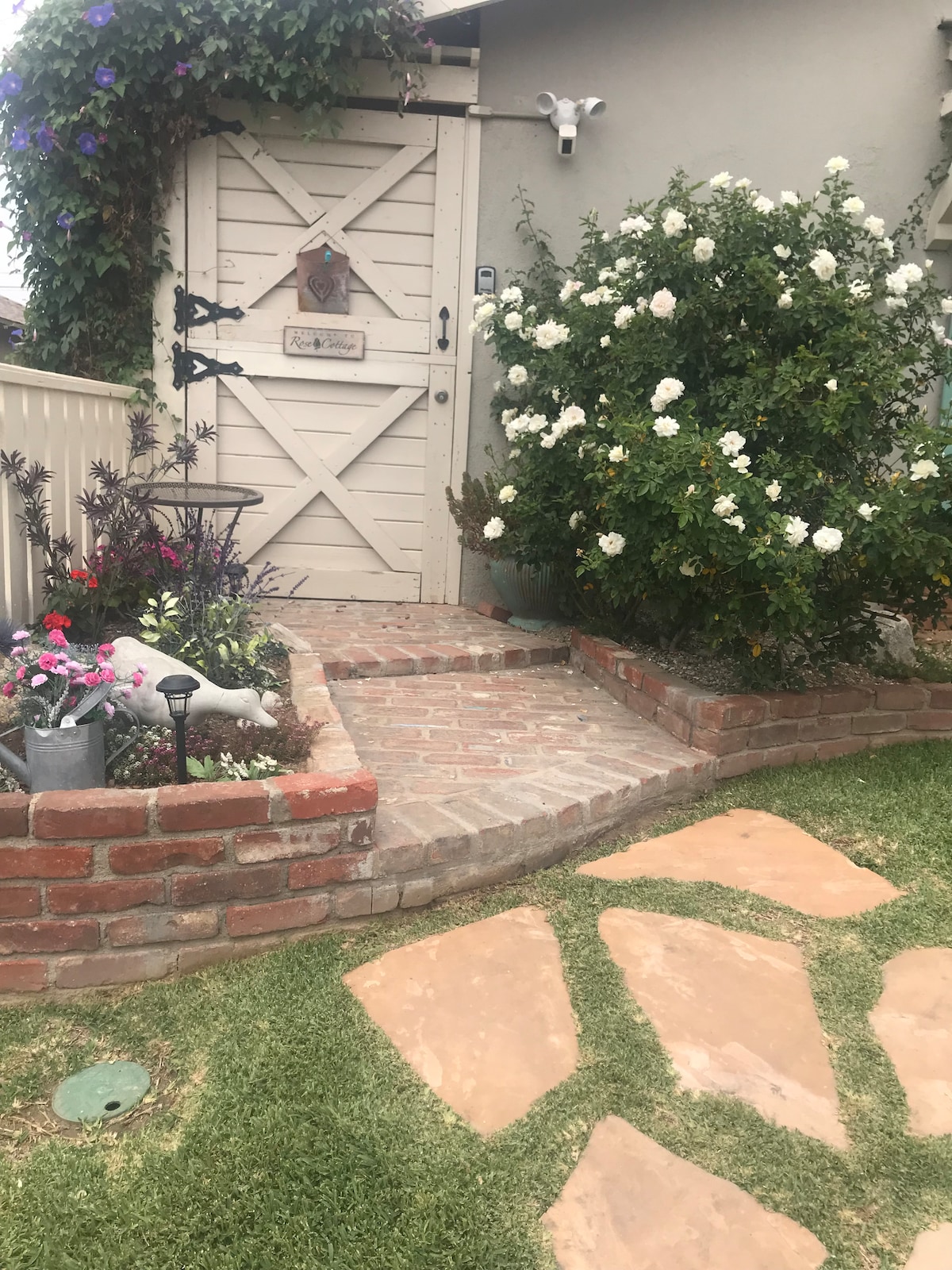 A charming entryway is adorned with lush greenery and colorful flowers leading up to a rustic wooden door. A brick pathway and decorative plant stands enhance the inviting atmosphere, while a white flowering bush adds a touch of natural beauty to the space.