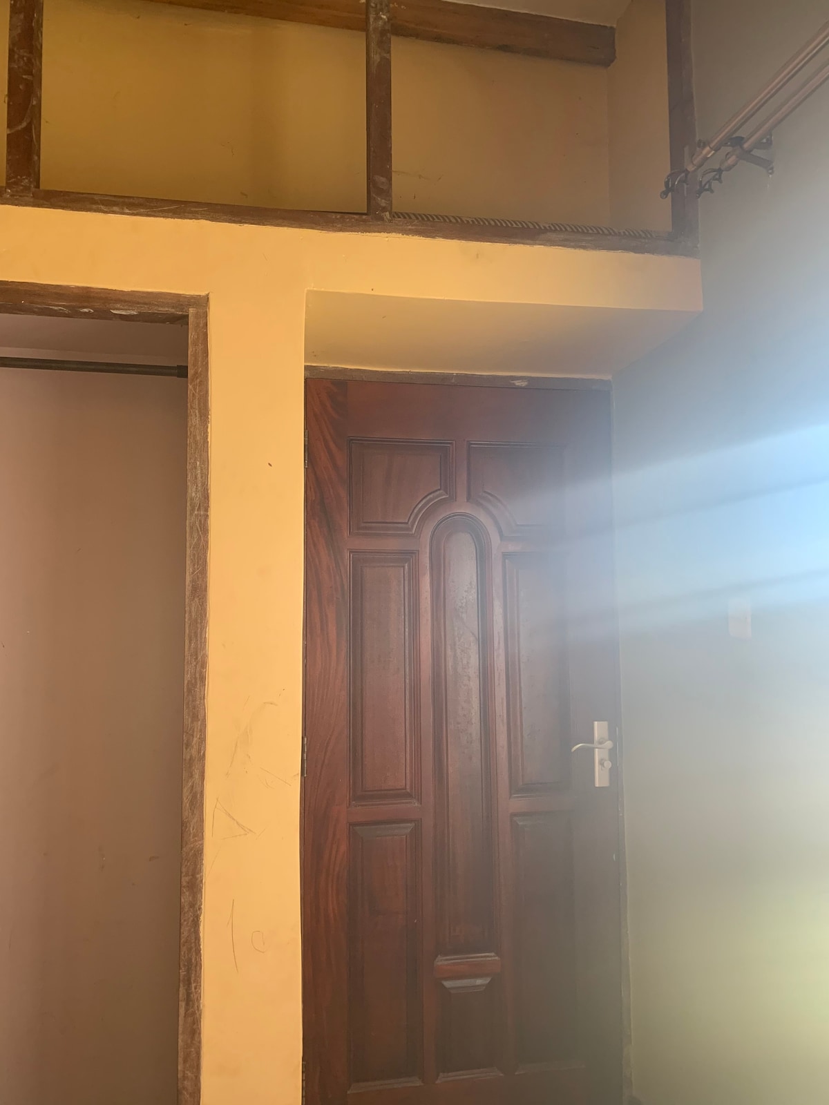 A wooden door with a decorative archway design stands as a focal point against softly lit yellow walls. Natural light streams through a nearby window, creating a gentle interplay of shadows. The adjacent wall appears muted, blending with the overall calm ambiance.